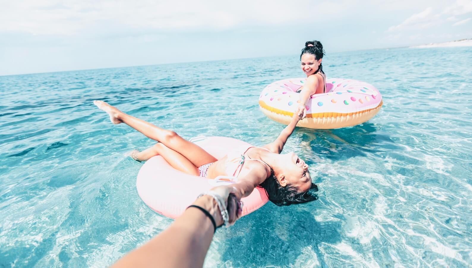 Two women laughing while floating on inflatable donuts in the ocean at the beach showcasing their Bubbly Personality.