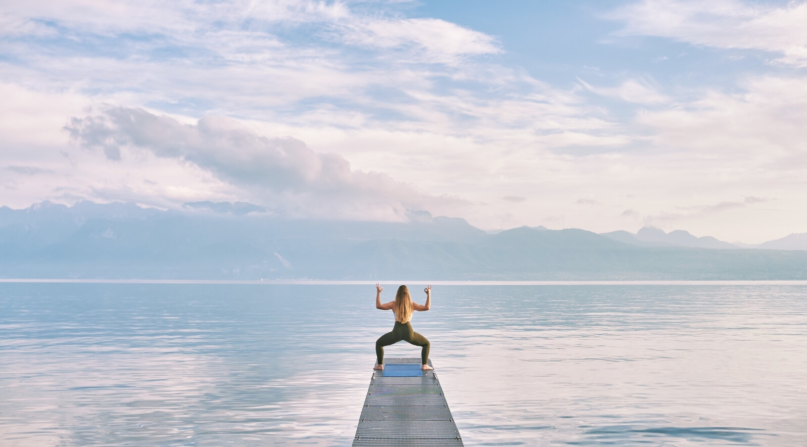 A woman meditating on a pier overlooking serene water with mountains in the background