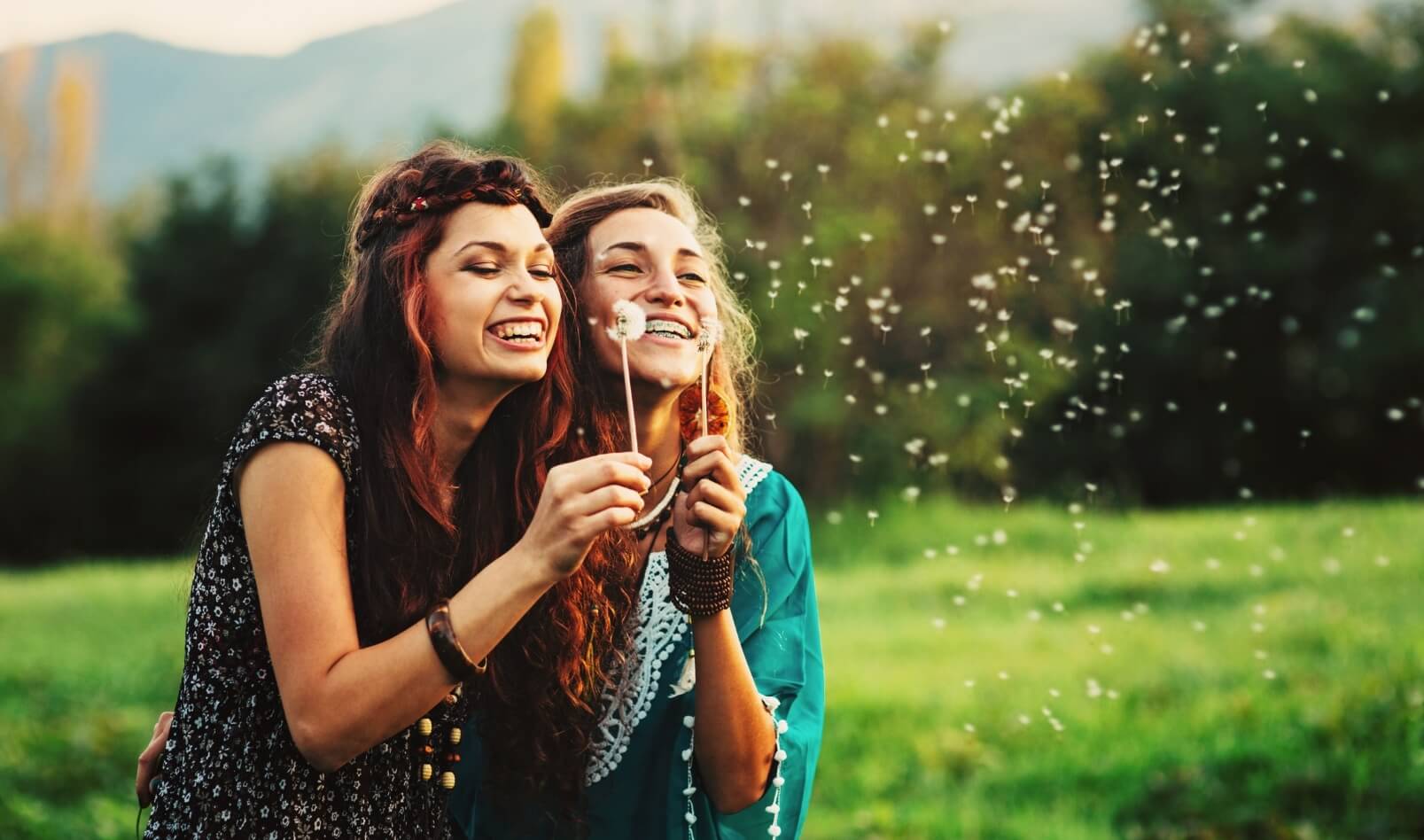 Two girls in a field releasing dandelion seeds from a dandelion flower.
