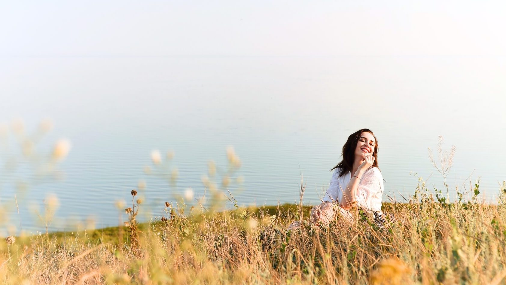 Woman practicing mindfulness for improved focus, sitting in a meadow with the ocean behind.