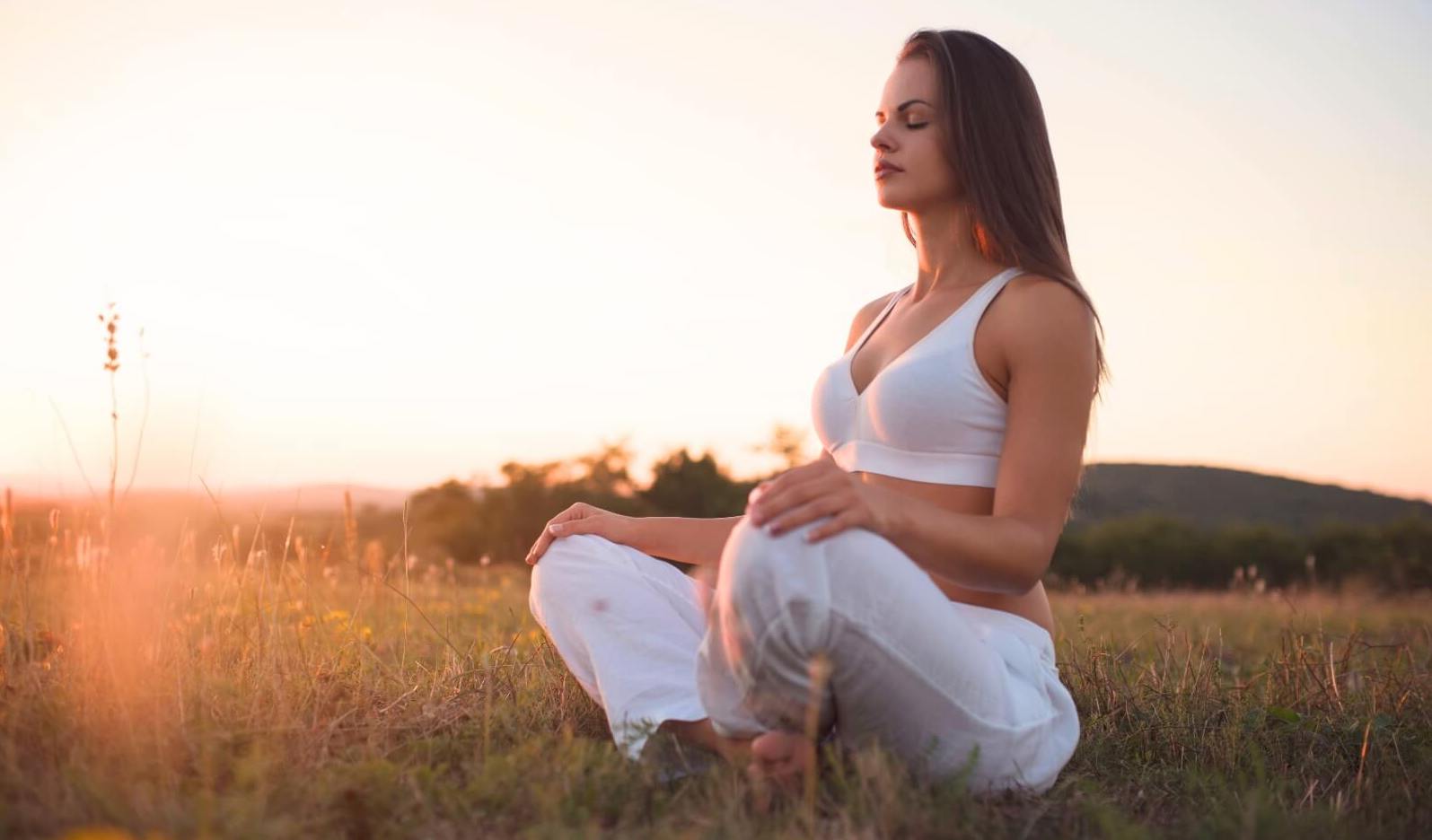 A woman with her legs crossed and eyes closed practicing mindfulness vs meditation in a meadow at sunset.