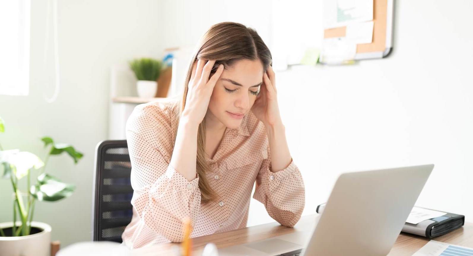 A woman sitting at her computer with her eyes closed and her hands on her head, showing signs of workplace stress.